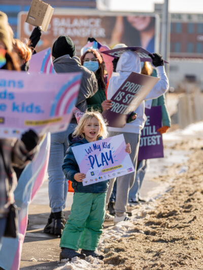 Image of a kid holding a sign that says, "Let trans kids play"
