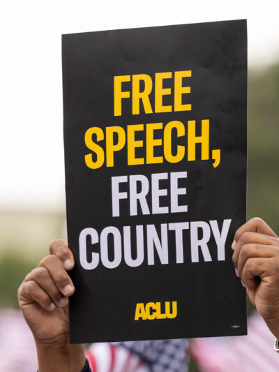 A demonstrator (with only their hands visible) holds up a sign that reads " FREE SPEECH, FREE COUNTRY - ACLU".