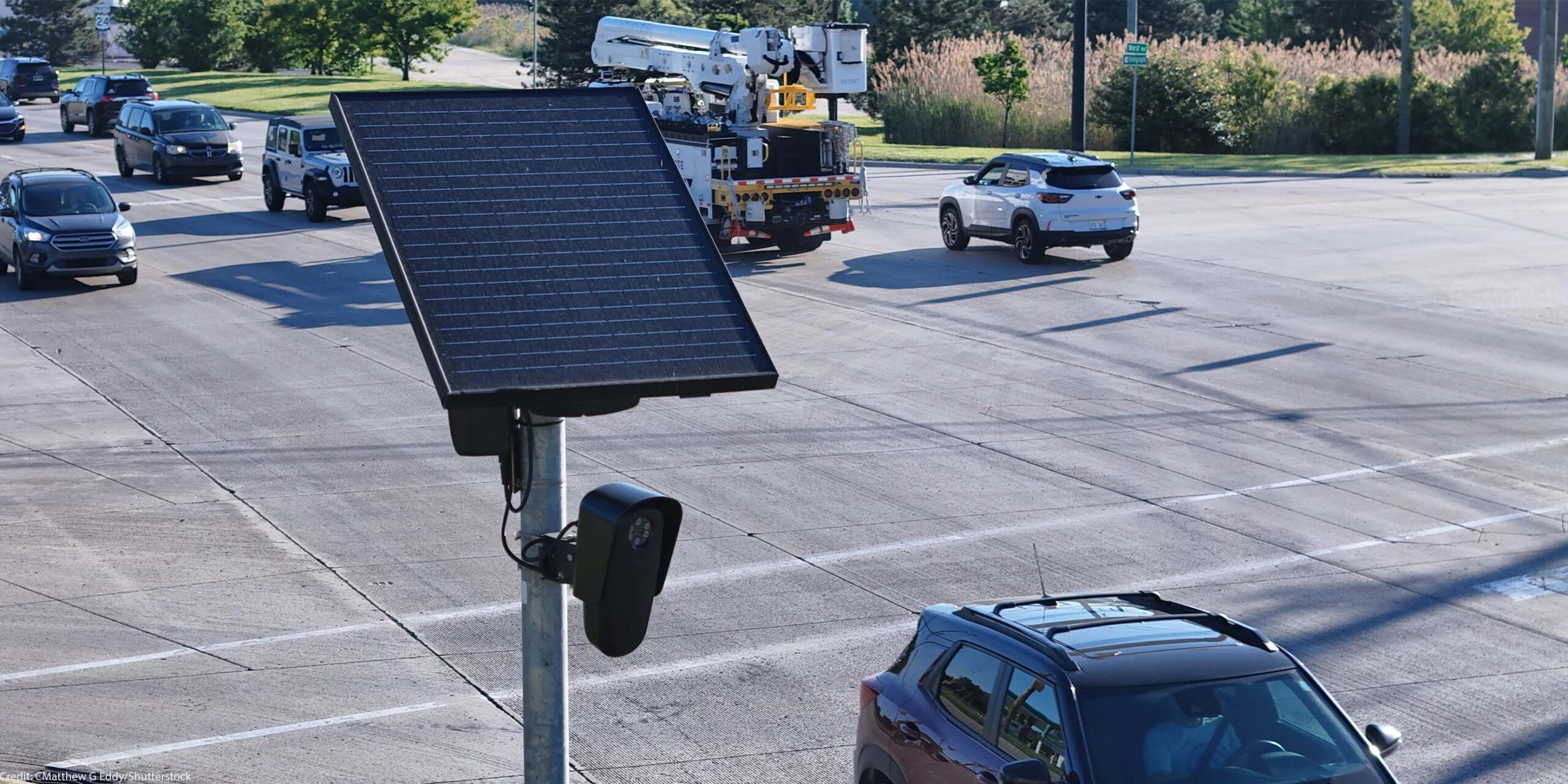 A solar-powered Flock camera is mounted on a pole beside a busy multi-lane road, with cars and a utility truck driving through the intersection in the background.
