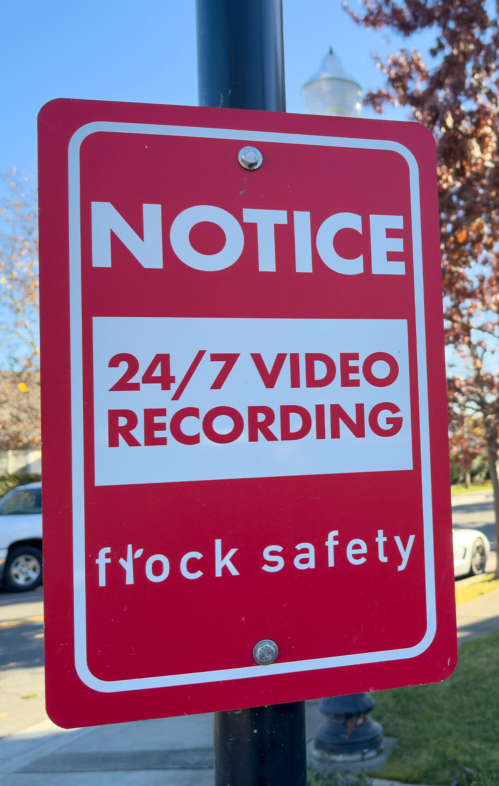 A red Flock Safety sign on a pole reads "Notice: 24/7 Video Recording" on a residential street, with parked cars, homes, trees, and a clear blue sky in the background.