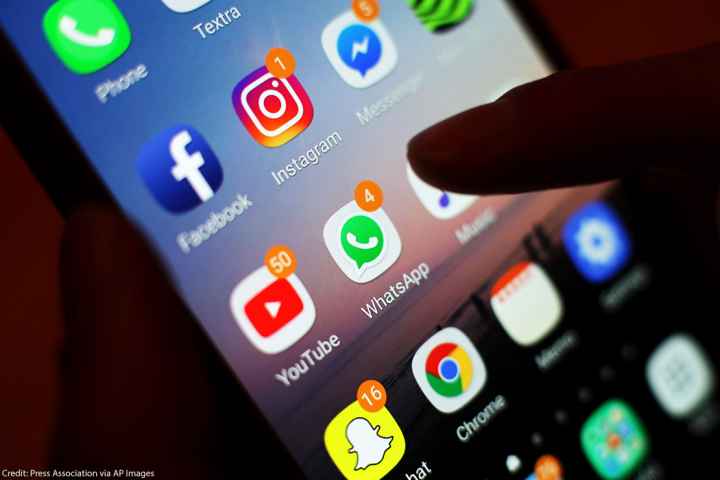 A hand holding a phone whose screen displays a group of apps, including Facebook, Instagram, and WhatsApp.