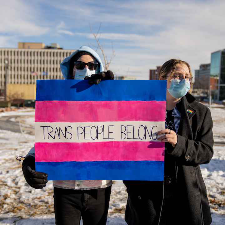 Image of two people holding a sign that reads, "trans people belong."